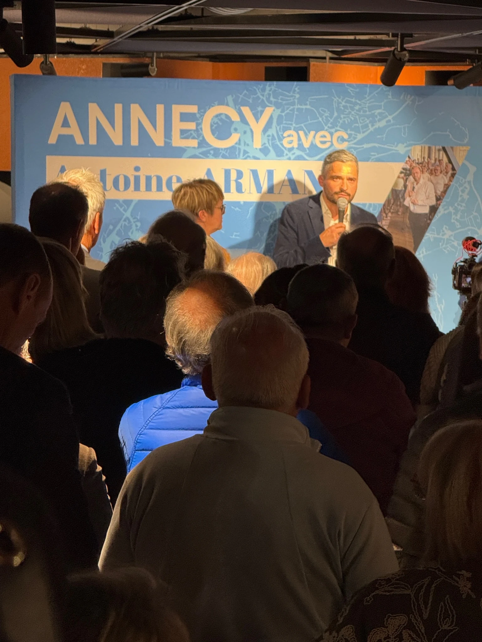 Assemblée publique avec un intervenant debout sur scène lors d'un meeting politique à Annecy, entouré d'un public nombreux - Réunion publique et concertation citoyenne - Acteurs Annecy