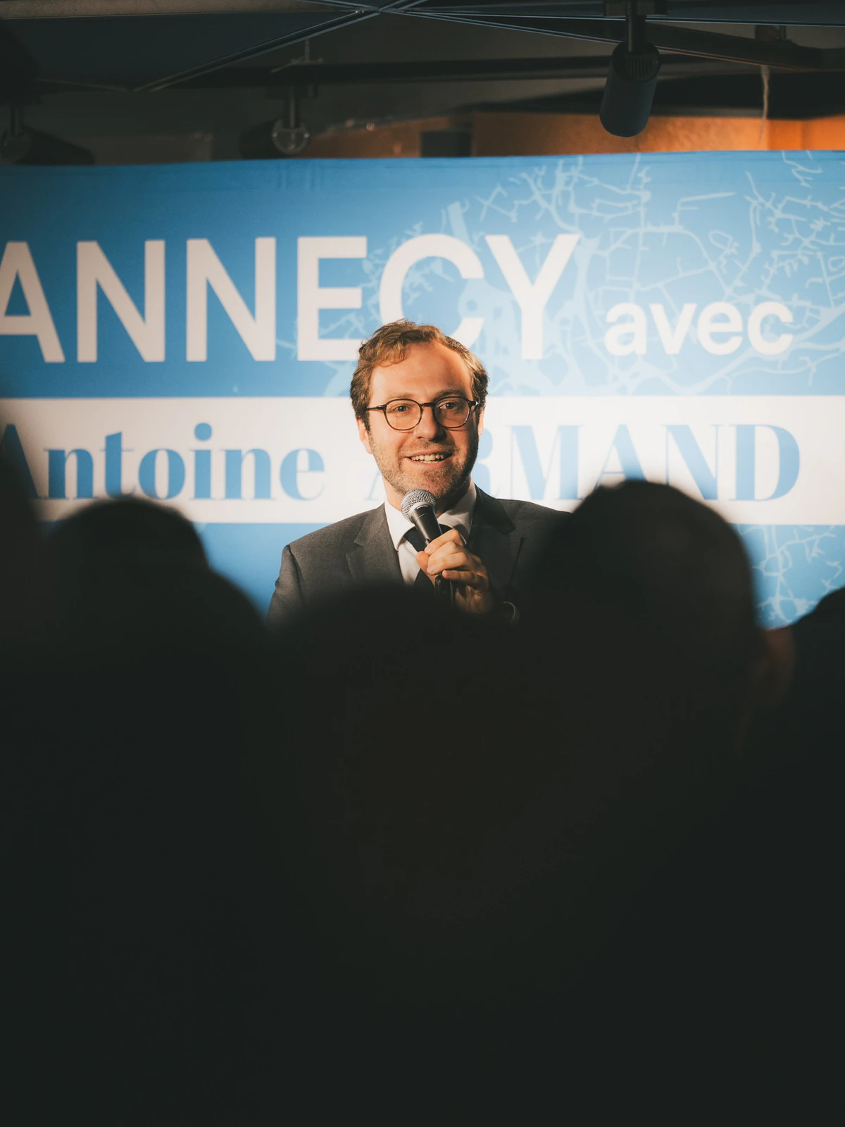Orateur à Annecy lors d'un événement politique avec bannière Antoine Armand et public en première ligne - Nos cinq engagements pour toutes les communes déléguées