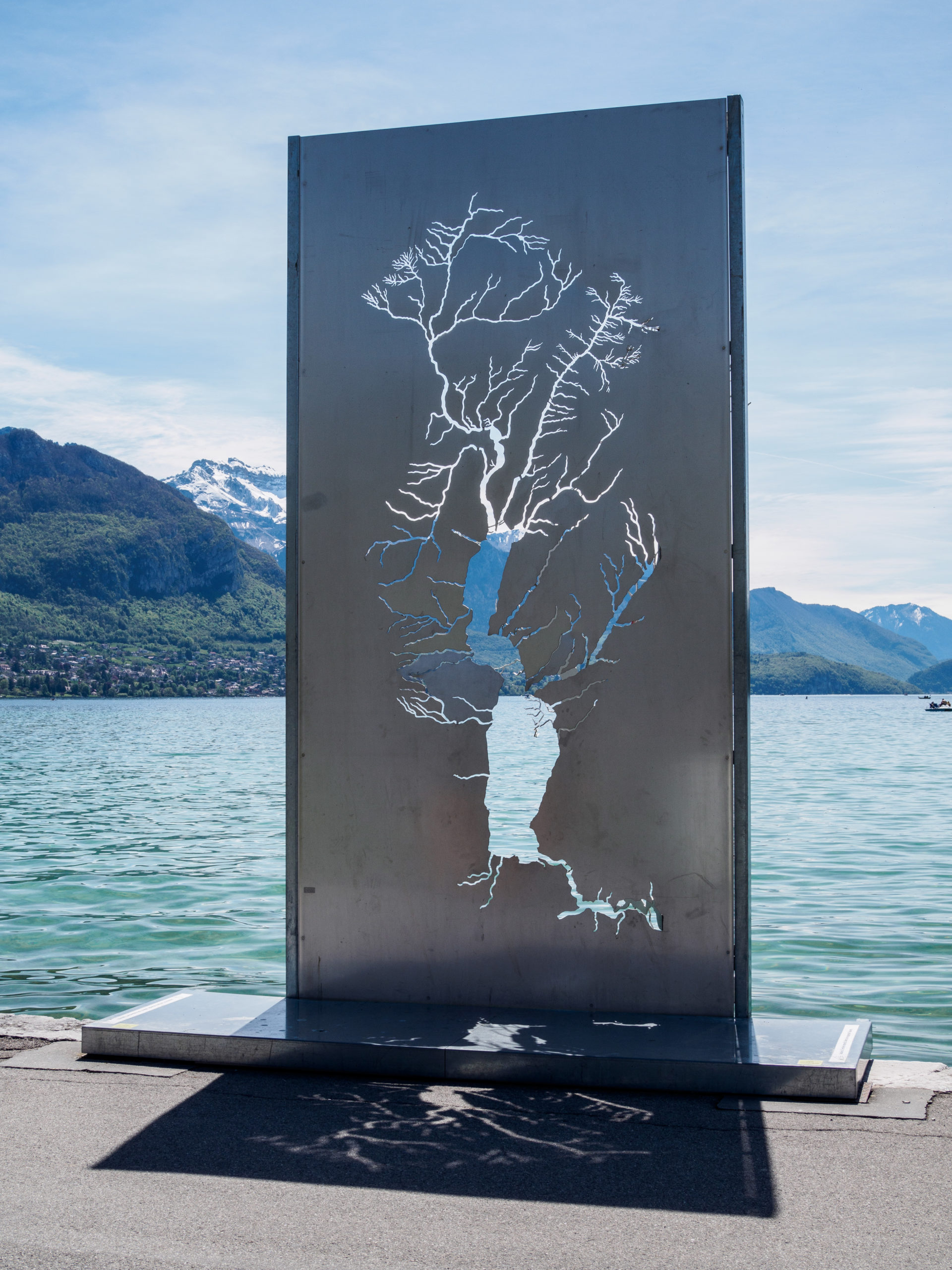 Œuvre d’art métalique à Annecy Sculpture métallique représentant le bassin versant du lac d’Annecy, découpée dans une plaque dressée au bord de l’eau, avec les montagnes en arrière-plan sous un ciel bleu clair. Photographie utilisée pour le site internet les Acteurs d'Annecy.