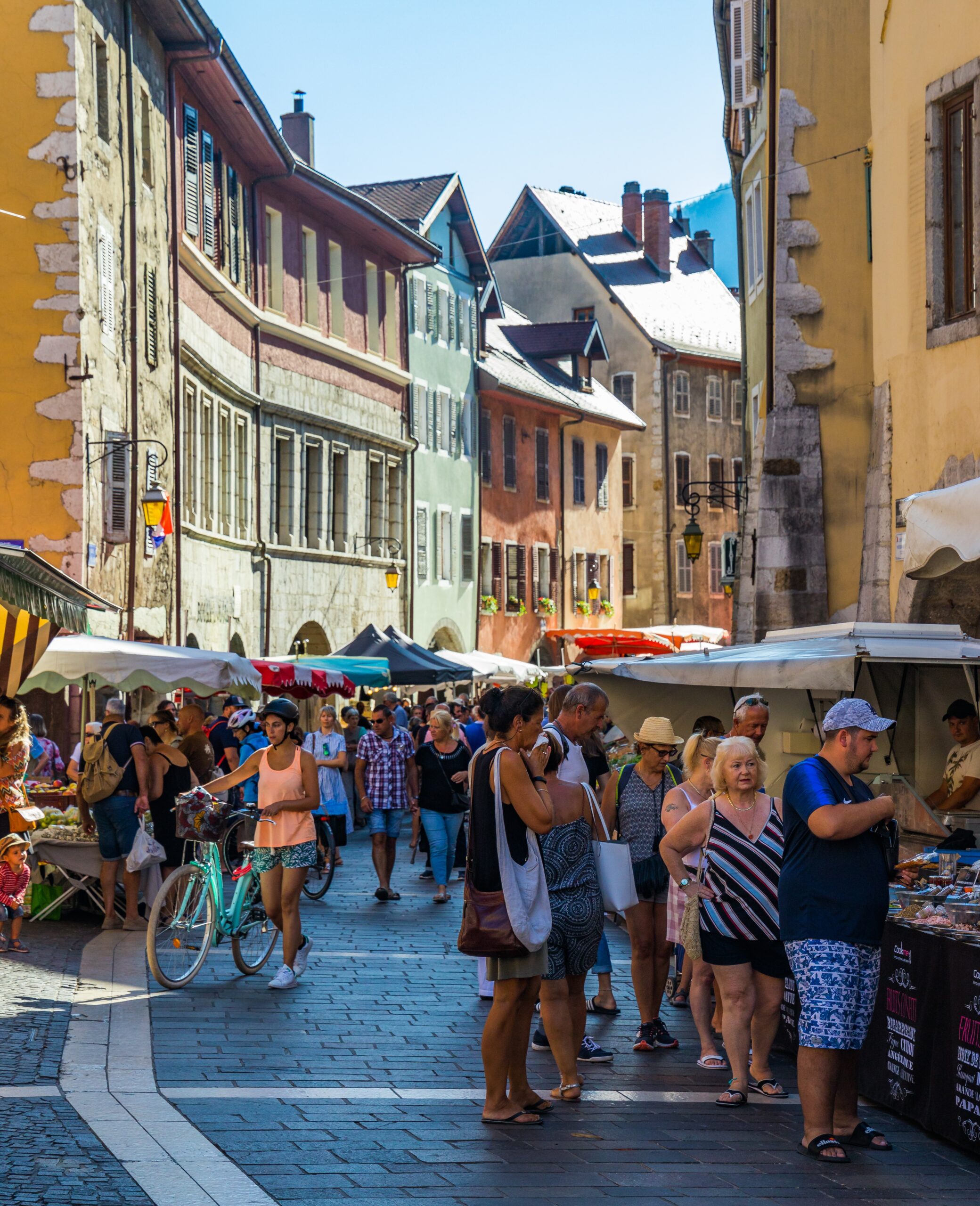 Marché animé dans les rues colorées de la vieille ville d’Annecy, avec de nombreux passants, stands de produits locaux et façades typiques savoyardes sous un ciel bleu d’été. Photographie utilisée pour le site internet les Acteurs d'Annecy.